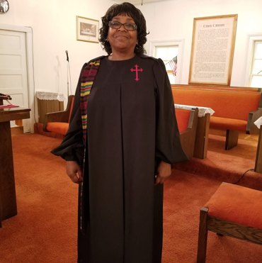 Elder Sandra Hodge in a black robe with a red cross, standing in the sanctuary